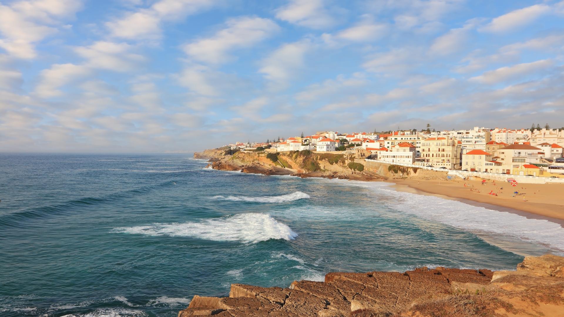Blick von der Küste aus auf das Meer und die kleine Sadt Sintra mit Strand und vielen weißen Häusern und orangenen Dächern.