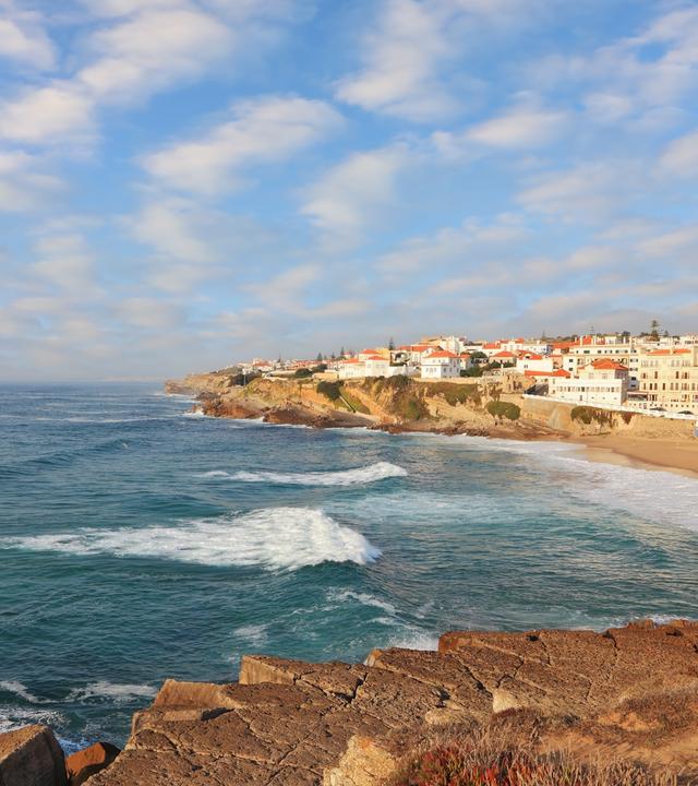Blick von der Küste aus auf das Meer und die kleine Sadt Sintra mit Strand und vielen weißen Häusern und orangenen Dächern.