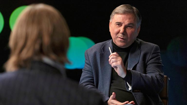 van Krastev schaut Richard David Precht an und hält eine Hand nach oben. Vorne ist Richard David Precht sitzend von hinten in einer Halbtotale unscharf zu sehen.
