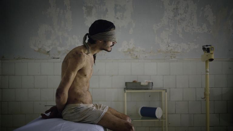"La noche de 12 años":  “Ruso” Rosencof (Chino Darín Mauricio) sitzt mit nacktem Oberkörper, auf dem Rücken zusammengebundenen Händen und verbundenen Augen auf einer Pritsche in einem heruntergekommenen Untersuchungsraum.