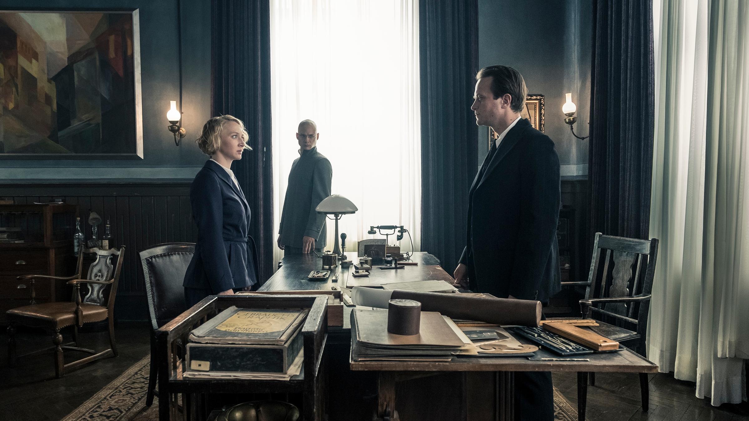 "Die neue Zeit":  Dörte (Anna Maria Mühe) und Gropius (August Diehl) stehen sich an dessen Schreibtisch gegenüber. Im Hintergrund steht Itten am Fenster, der von der Affäre zwischen ihnen weiß.
