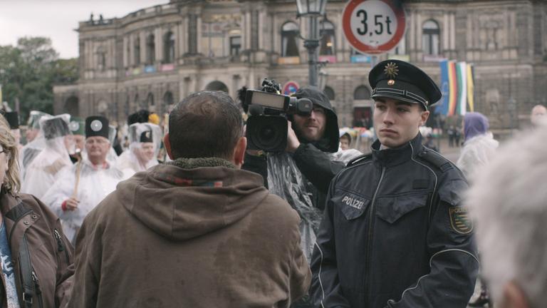 "AGGREGAT": Ein Pegida-Anhänger beschimpft vor der Semperoper einen Kameramann und wird von einem Polizisten dabei beobachtet.