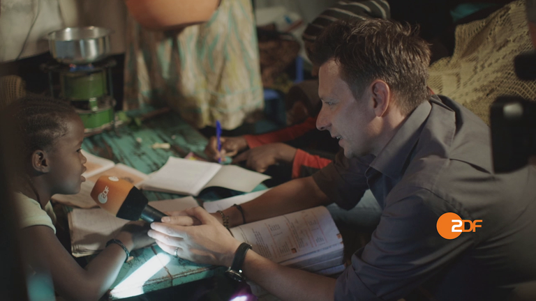 Auslandskorrespondenten-Kampagne: Timm Kröger interviewt ein Schulkind in Afrika, dass an einem Tisch mit ausgebreiteten Arbeitsuntensilien sitzt.