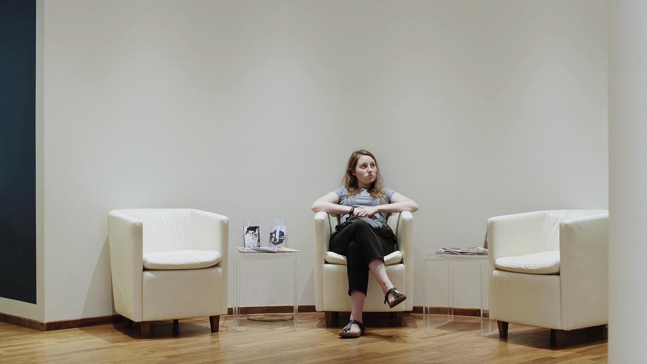 "Ab 18! - Dazwischen Elsa": Elsa sitzt mit übereinander geschlagenen Beinen in einem weißen Ledersessel vor einer Wand und schaut nachdenklich. Links und rechts von ihr jeweils ein Beistelltisch und ein leerer Sessel.