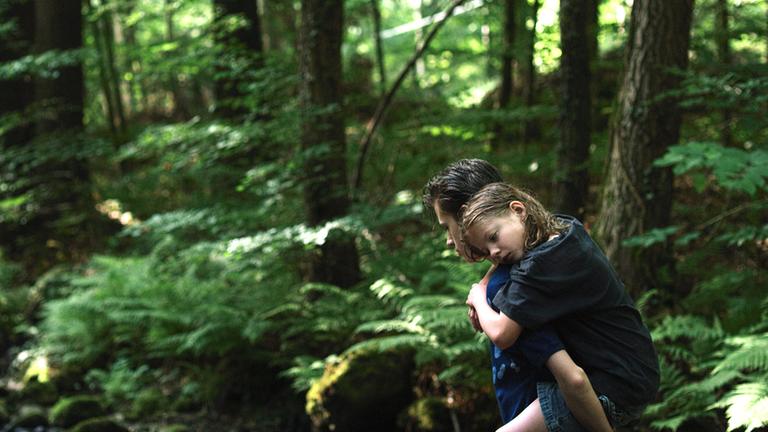 "Ich war zuhause, aber" (3sat): Phillip (Jakob Lassalle) läuft mit seiner kleinen Schwester Flo (Clara Möller) auf dem Rücken durch den Wald.