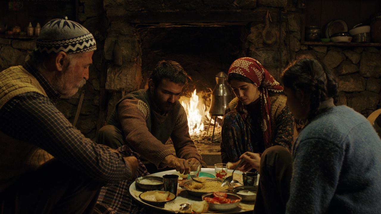 "Eine Geschichte von drei Schwestern": Veysel (Kayhan Açıkgöz),  Şevket (Müfit Kayacan),  Reyhan(Cemre Ebüzziya),  Havva(Helin Kandemir) sitzen beim Essen am Tisch. Im Hintergrund brennt ein Feuer im offenen Kamin..