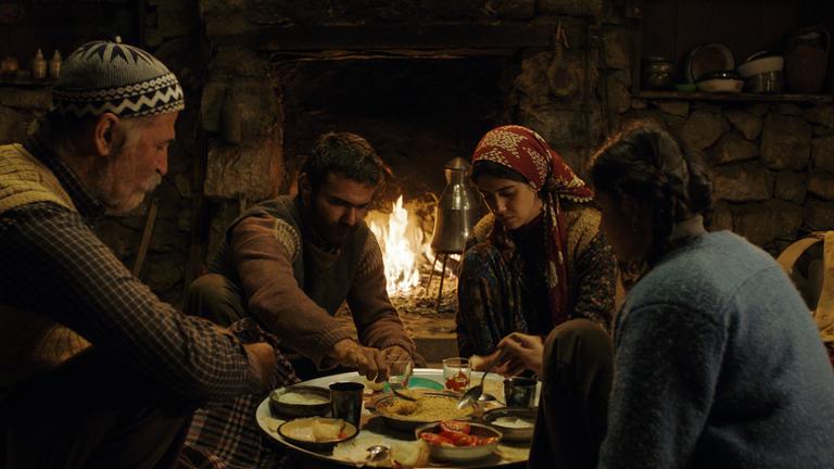 "Eine Geschichte von drei Schwestern": Veysel (Kayhan Açıkgöz),  Şevket (Müfit Kayacan),  Reyhan(Cemre Ebüzziya),  Havva(Helin Kandemir) sitzen beim Essen am Tisch. Im Hintergrund brennt ein Feuer im offenen Kamin..