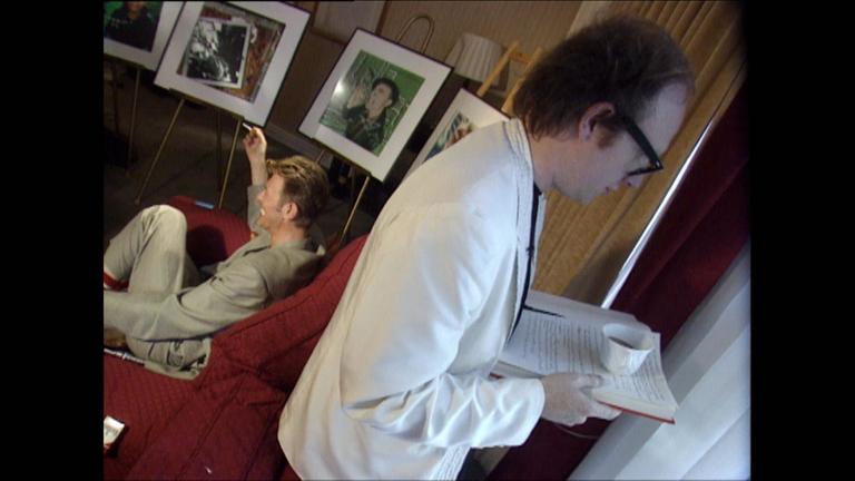 "Why are we creative": Mann auf dem Sofa sitzend guckt nach links, anderer Mann steht rechts im Bild mit Buch in der Hand mit dem Rücken zum Gesprächspartner.