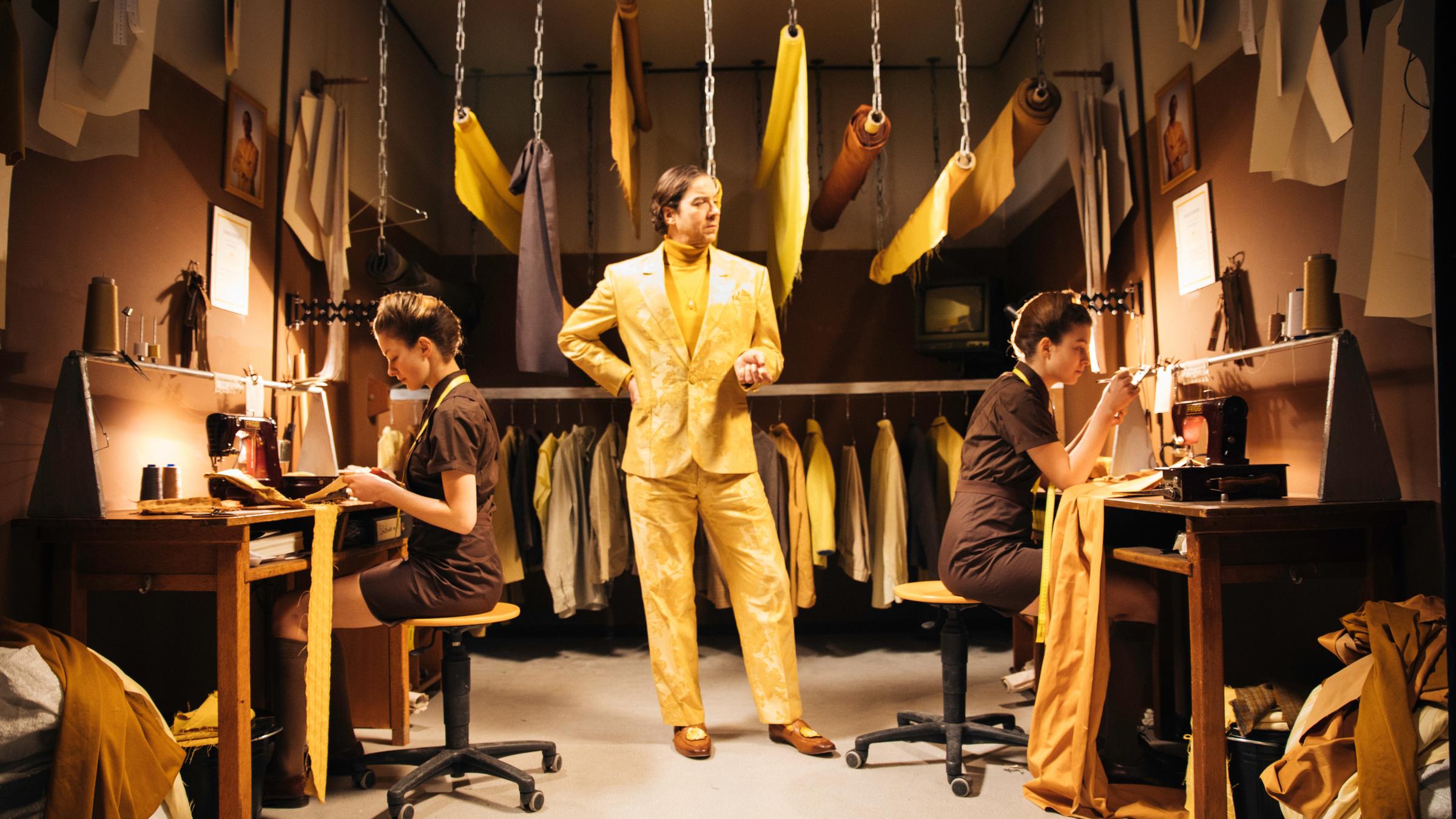 KlassiXS - die großen Dramen in jungen Kurzfilmen - Kaiser: Blick in eine Schneiderei. Im Hintergrund hängen Stoffe von der Decke und Kleidung am Haken. Rechts und links an der Wand sind Arbeitsplätze mit Nähmaschinen, an jedem Arbeitsplatz sitzt eine Frau in einem braunen Kostüm und arbeitet. Die beiden Frauen sind Zwillinge. In der Mitte des Raums steht ein Mann in einem gelben Anzug und schaut den Schneiderinnen bei der Arbeit zu.