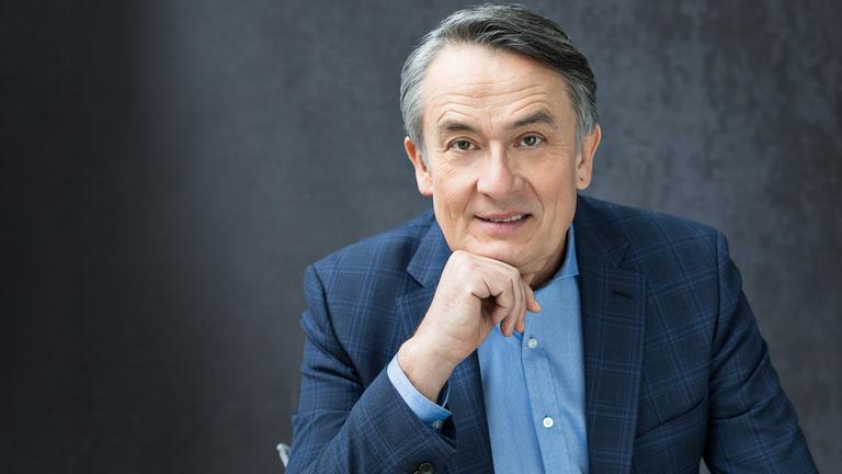 "Gert Scobel": Porträtbild von Gert Scobel (nur Oberkörper). Er trägt ein hellblaues Hemd und ein blaukariertes Sakko. Er schaut lächelnd in die Kamera