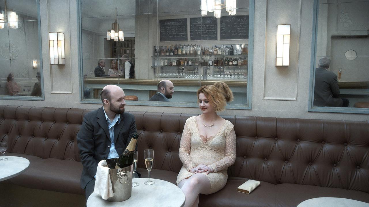 "Über die Unendlichkeit" (ARTE): Ein Mann (Pär Frederiksson) und eine Frau (Lisa Blohm) sitzen auf einem Ledersofa in einer Bar. Sie sind festlich gekleidet. Auf dem Tisch steht ein Sektkübel min einer Flasche und zwei Gläsern. Im Hintergrund der Bartresen.