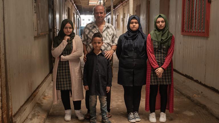 Kein Recht auf Familie? Geflüchtete kämpfen um ihre Angehörigen (ARTE): Fünf Mitglieder einer Flüchtlingsfamilie stehen in einer Notunterkunft zwischen zwei Baucontainer-Reihen.