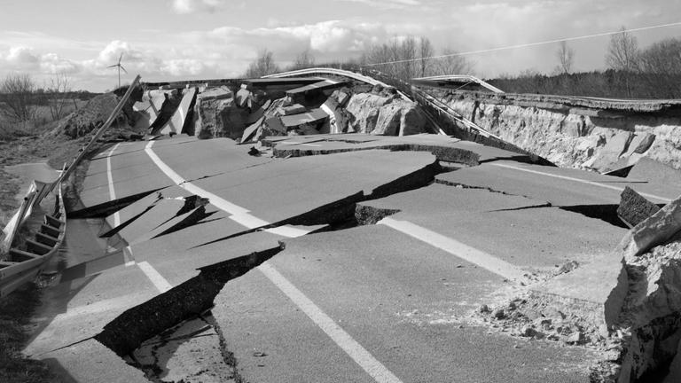 Schwarzweißes Landschaftsbild von einem Autobahnteilstück, dass durch Erosion eingebrochen ist, so dass Asphaltstücke sich aufwerfen und tiefe Risse und Spalten entstanden sind.