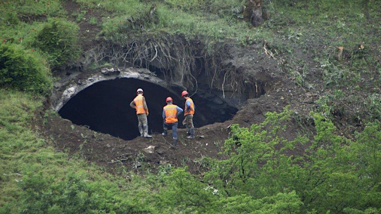 "Der Fluch des Tunnels": Drei Männer in Arbeitskleidung stehen an einem Berghang und blicken in einen großen Tunneleingang