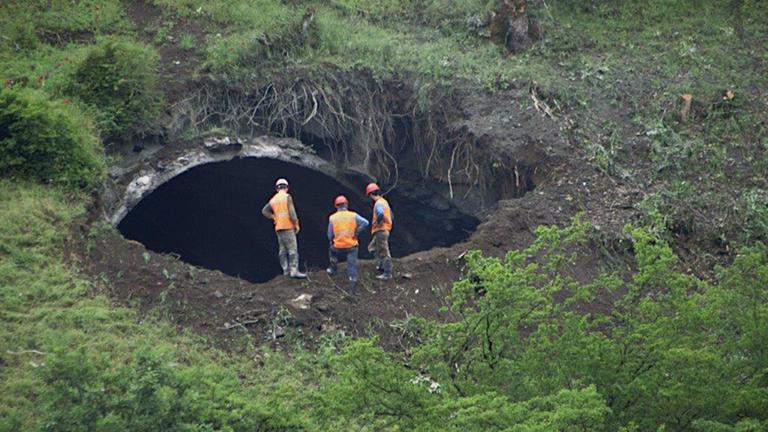 "Der Fluch des Tunnels": Drei Männer in Arbeitskleidung stehen an einem Berghang und blicken in einen großen Tunneleingang