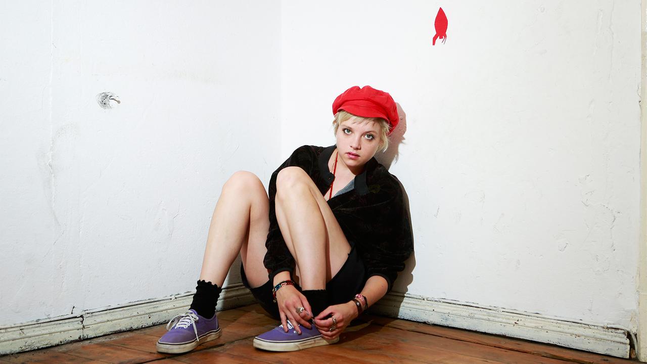 "Kokon": Romy (Jella Haase) sitzt in einem kahlen Raum an die Wand geleht auf dem Boden und schhaut in die Kamera.