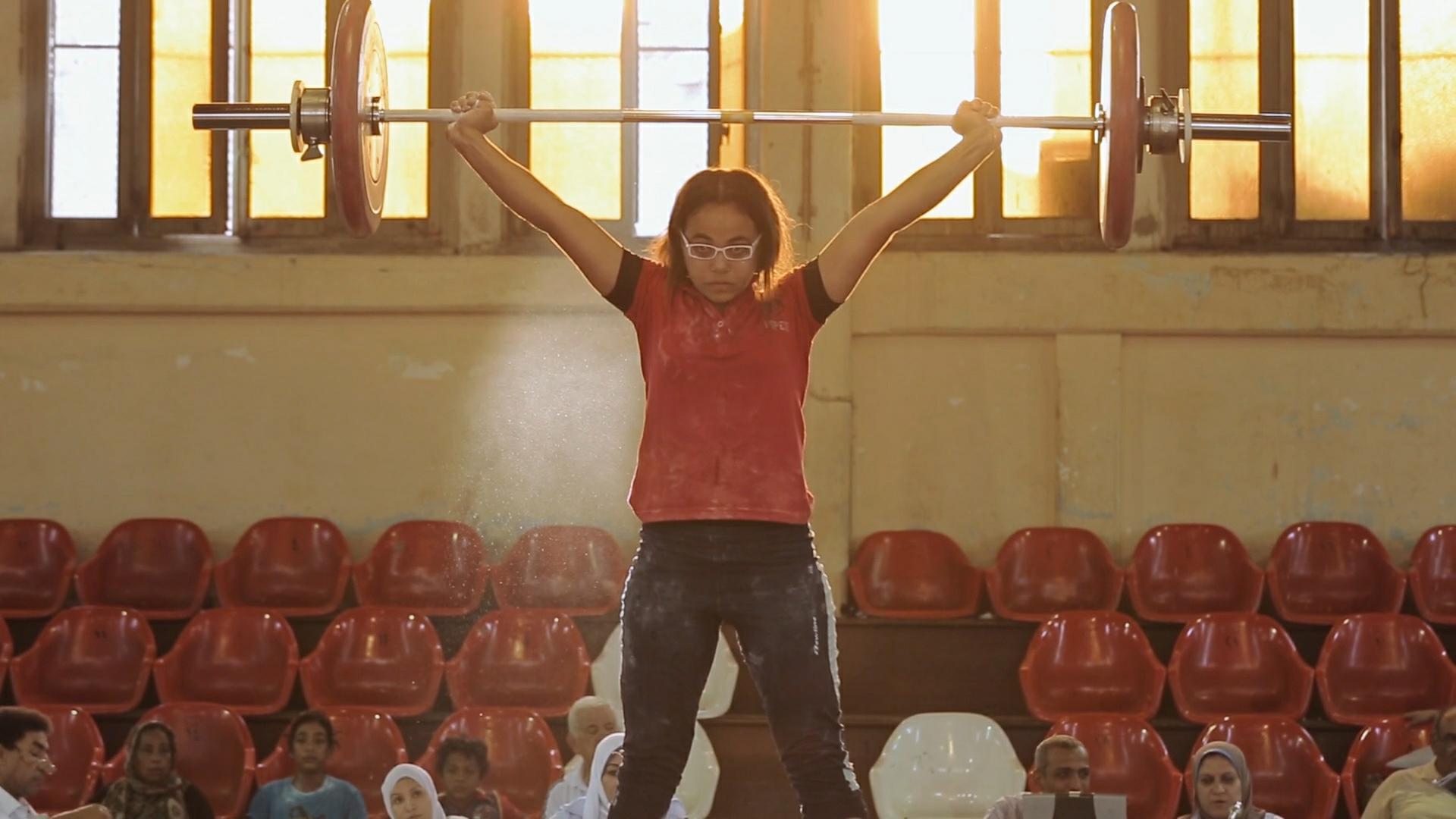 "Lift Like a Girl - Stark wie ein Mädchen": Zebiba stemmt ein Gewicht beim Nationalen Jugendturnier in Ägypten. Im Hintergrund Zuschauerränge.