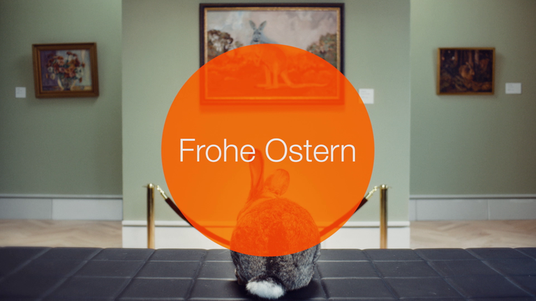 Screenshot Oster-Spots 2020 - Falsche Hasen. Ein Hase, überdeckt von einem orangenen Kreis mit den Worten "Frohe Ostern"  schaut sich in einem Museum Gemälde an, auf denen Hasen zu sehen sind. 