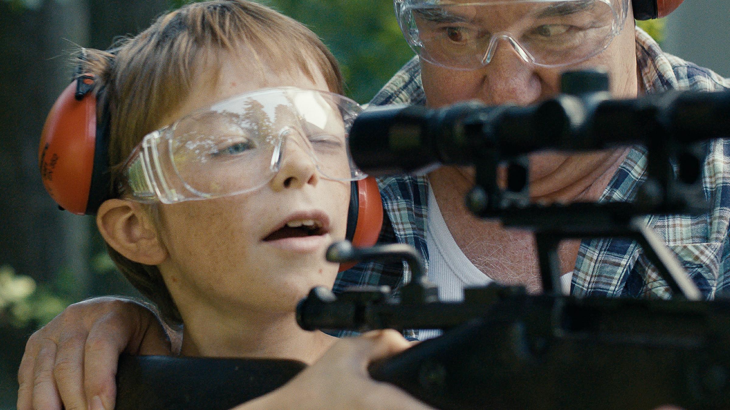 "Rivale": Roman (Yelizar Nazarenko) blickt durch das Zielfernrohr eines Gewehrs. Er trägt eine Schutzbrillle und Ohrenschützer und wird von einem älteren Mann links neben ihm assistiert