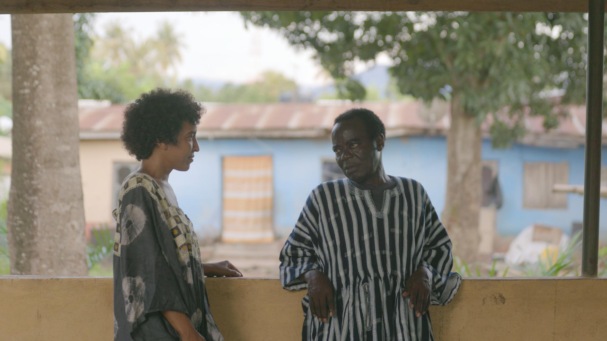 "König Bansah und seine Tochter": Katharina Bansah und ihr Vater Céphas Bansah (r.) in Ghana. Sie stehen uunter Bäumen vor einwem flachen Hasus und schauen sich an.