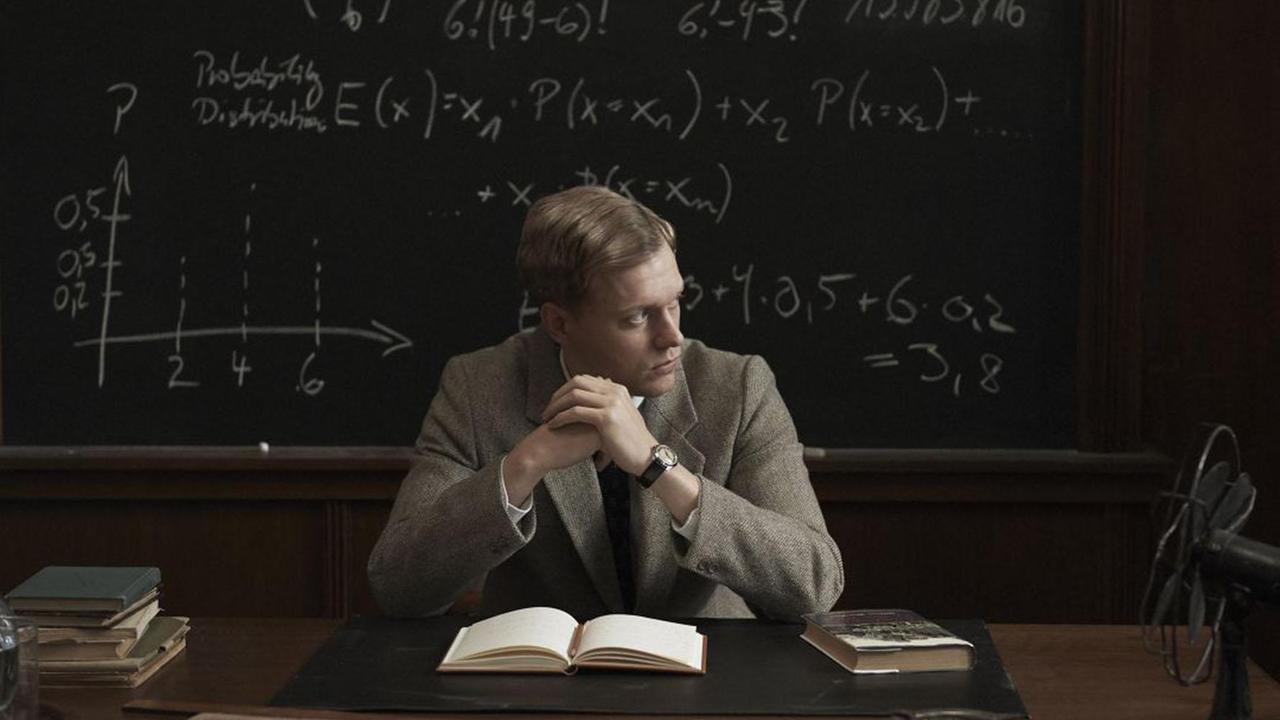 "Adventures of a Mathematician": Stan Ulam (Philippe Tlokinski)  sitzt an einem Pult vor einem aufgeschlagernern Buch und blickt zur Seite. Hinter ihm an der Wand eine Schultafwewl, auf die mathematische Formeln geschrieben sind. 