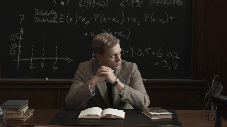 "Adventures of a Mathematician": Stan Ulam (Philippe Tlokinski)  sitzt an einem Pult vor einem aufgeschlagernern Buch und blickt zur Seite. Hinter ihm an der Wand eine Schultafwewl, auf die mathematische Formeln geschrieben sind. 