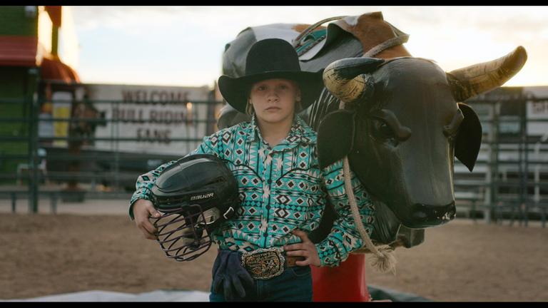 "Glitzer und Stau": Eine junge Rodeo-Reiterin mit Cowboyhut posiert neben einem Bullriding-Simulator. Unter ihren rechten Arm hat sie einen Schutzhelm geklemmt.