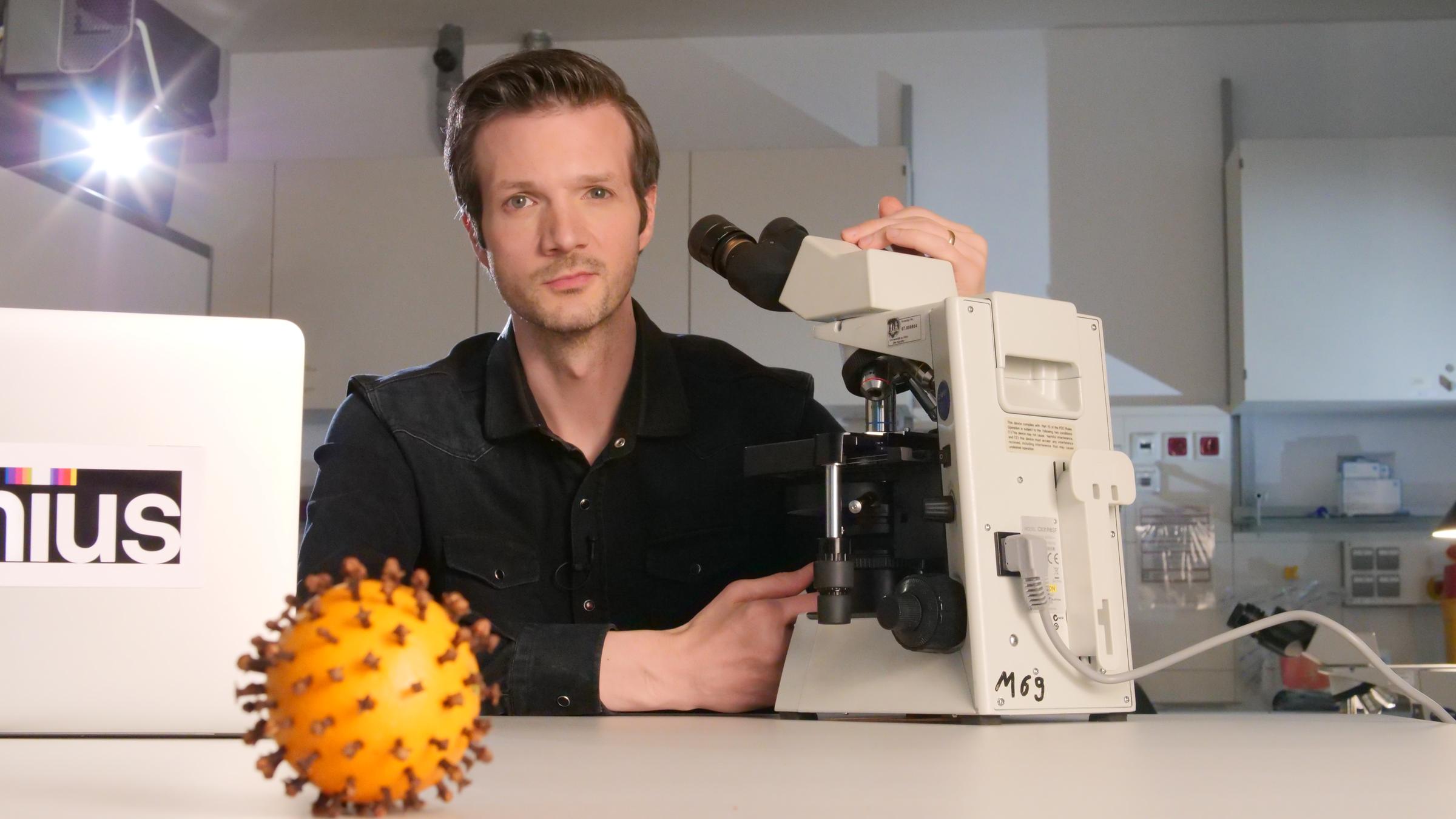 "Xenius: Zoonosen - Wie das Coronavirus den Menschen befiel" (ARTE): Xenius-Moderator Adrian Pflug untersucht Bakterien unter einem Lichtmikroskop – Viren allerdings sind dafür zu klein. Im Vordergrund: Eine mit Nelken gespickte Orange als symbolisches SARS CoV 2 Virus.
