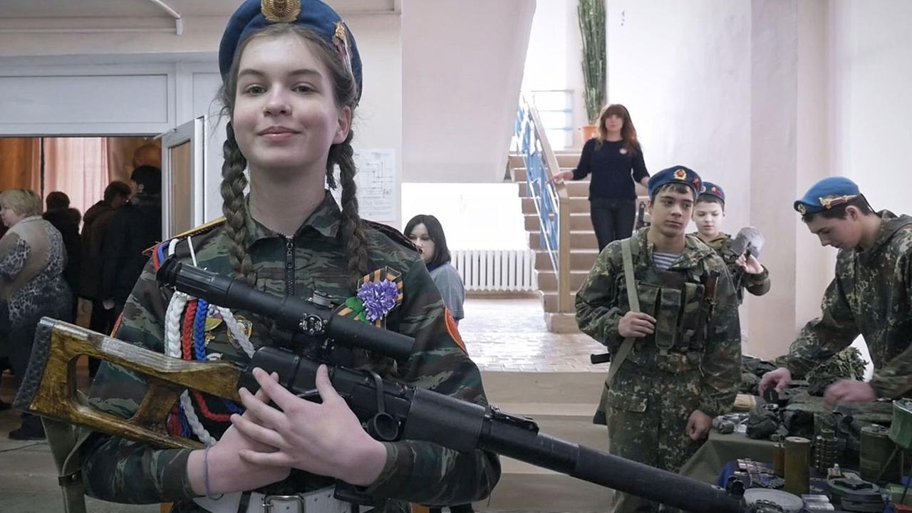 "Jelnja - Stadt des Ruhms" (ARTE): Ein jugendliches Mädchen in Soldatenuniform präsentiert auf einerWaffenschau mit stolzem Blick ein Maschinengewehr.  Im Hintergrund ddrei weitere, männliche Jugendliche in Soldatenuniformen an einem Tisch, auf dem Waffen liegen. 