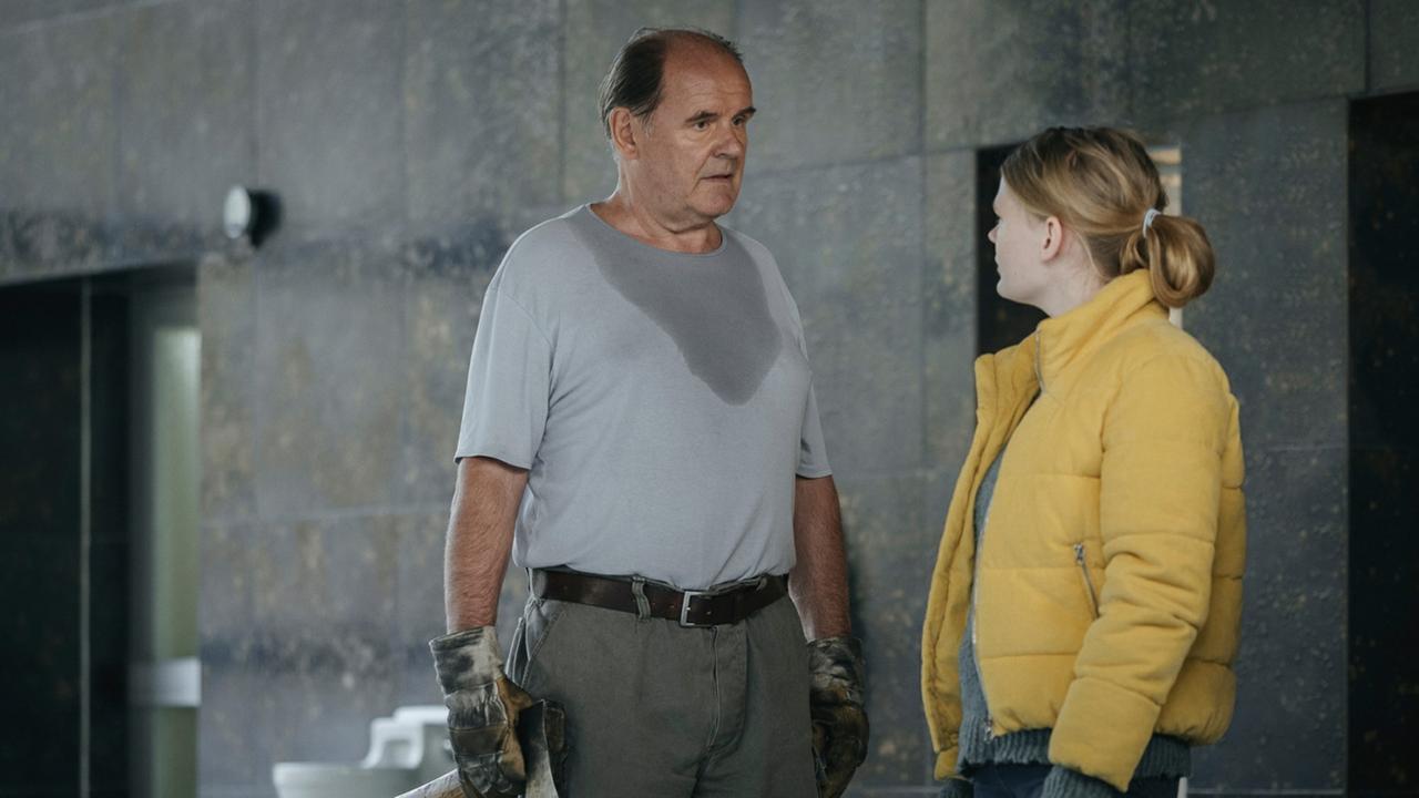 "Schlaf": Ein älterer Mann in einem verschwitzten T-Shirt, Arbeitshandschuhen und einem Beil in der rechtern Hand und eine jüngere Frau in einem gelben Anorak unterhalten sich auf einem düsteren Hotelflur 