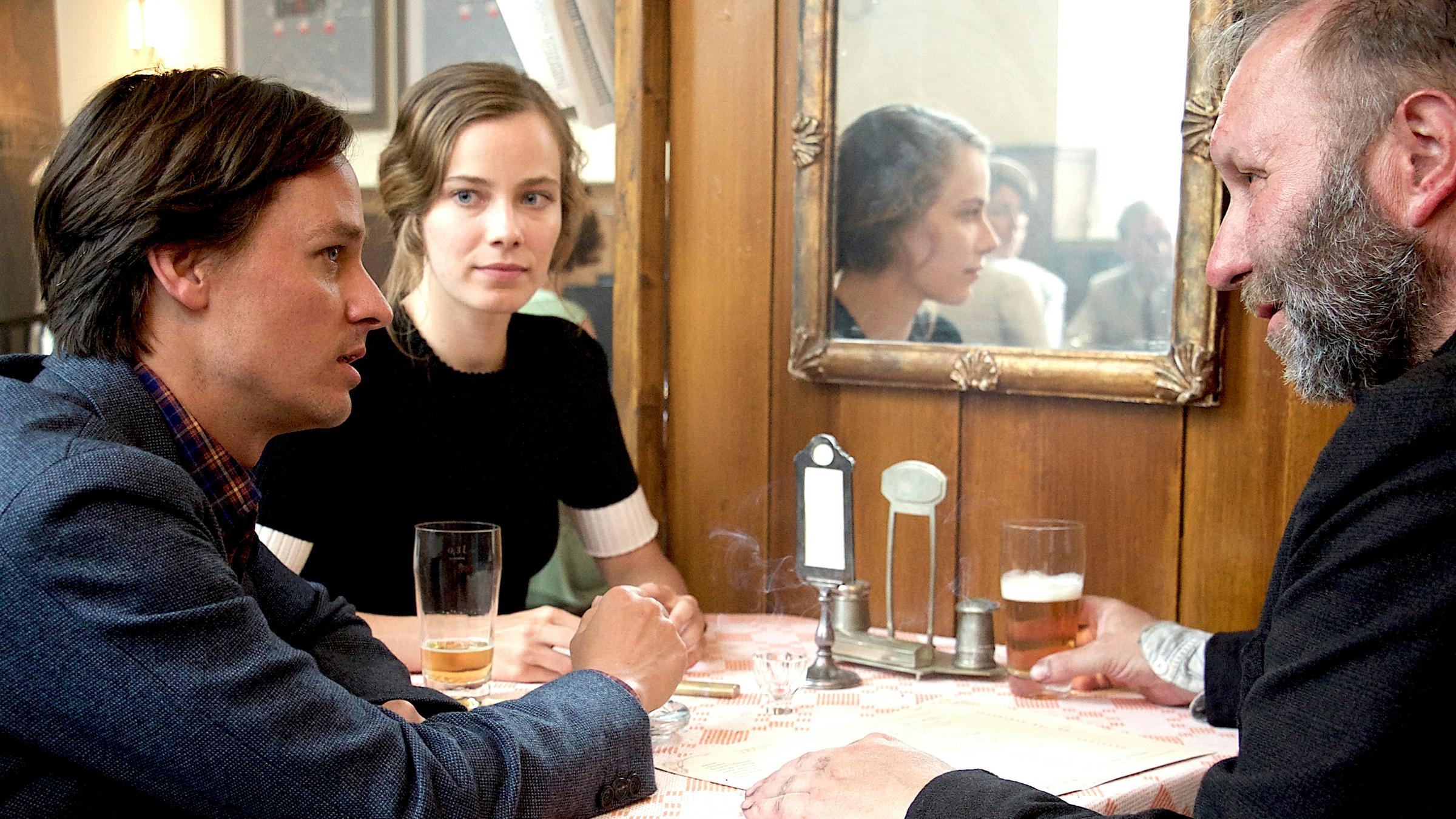 "Fabian oder der Gang vor die Hunde": Fabian (Tom Schilling, l.), Cornelia (Saskia Rosendahl) und Ray (Thomas Reimann) sitzen am Tisch in einer Gastwirtschaft und unterhalten sich.