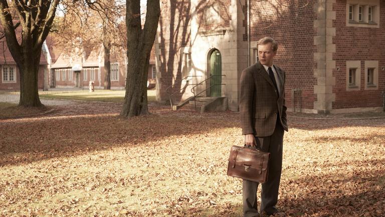 "Adventures of a Mathematician" (ARTE): Mathematiker Stan Ulam (Philippe Tlokinski) steht im Anzug und mit einer Aktentasche in der rechten Hand auf dem Rasen eines parkartigen Grundstücks und blickt skeptisch nach rechts. Im Hintergrund mehrere Backsteingebäude und Bäume.