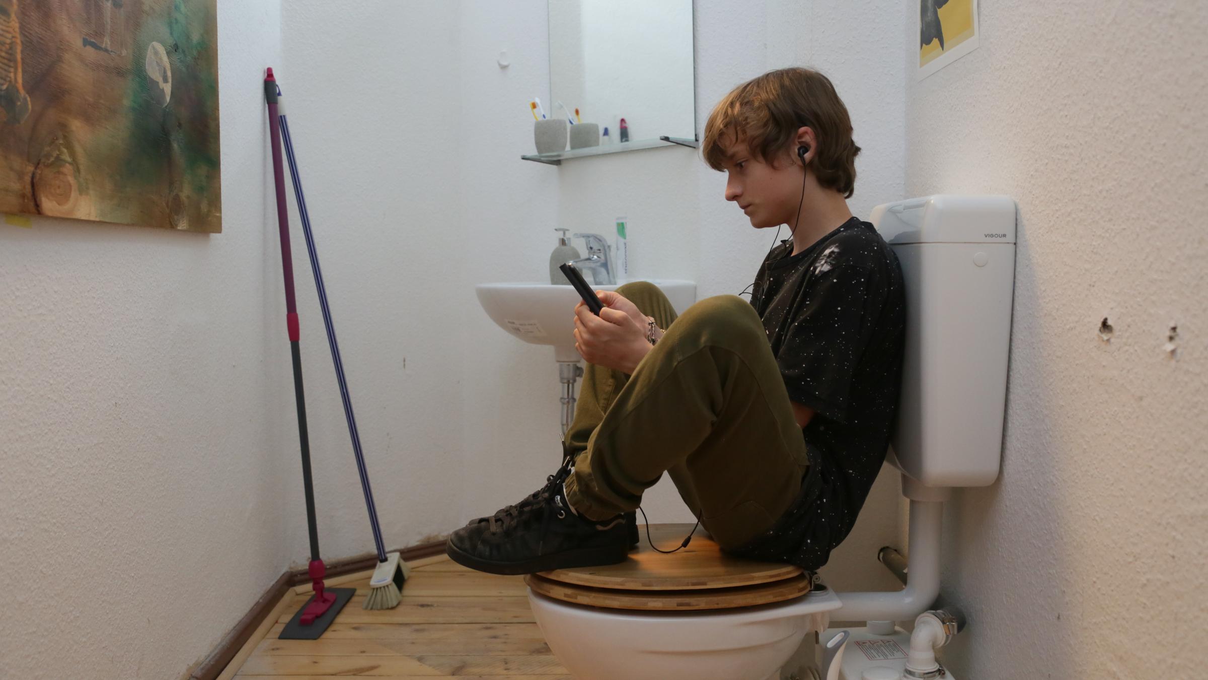  "@Kalinka08 - Melde dich bitte": Ein Junge sitzt auf der geschlossenen Toilette, die Beine hat er auf dem Toilettendeckel angewinkelt. In seinen Händen hält er ein Smartphone, in das er gespannt sieht. Er hat Kopfhörer in den Ohren.