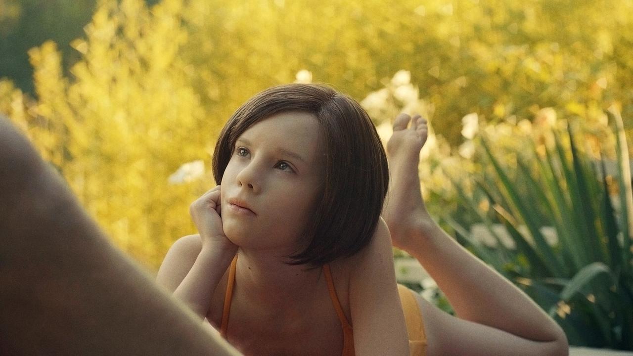 The Trouble with being born ("Die Not, geboren zu sein"): Android Elli (Lena Watson) liegt mit angewinkelten Beinen auf dem Bauch entspannt im Gras, das Kinn auf die rechte Hand gestützt und blickt zu einer Person vor ihr auf (nicht im Bild)