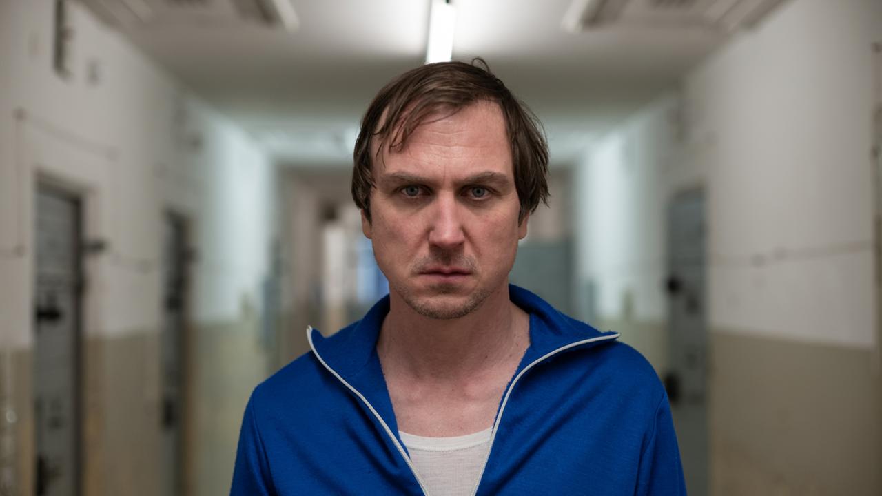 "Nahschuss": Franz Walter (Lars Eidinger) steht in einem blauen Trainingsanzug in einem Gefängnisgang und blickt frontal mit grimmigem Gersichtsausdruck in die Kamera. 