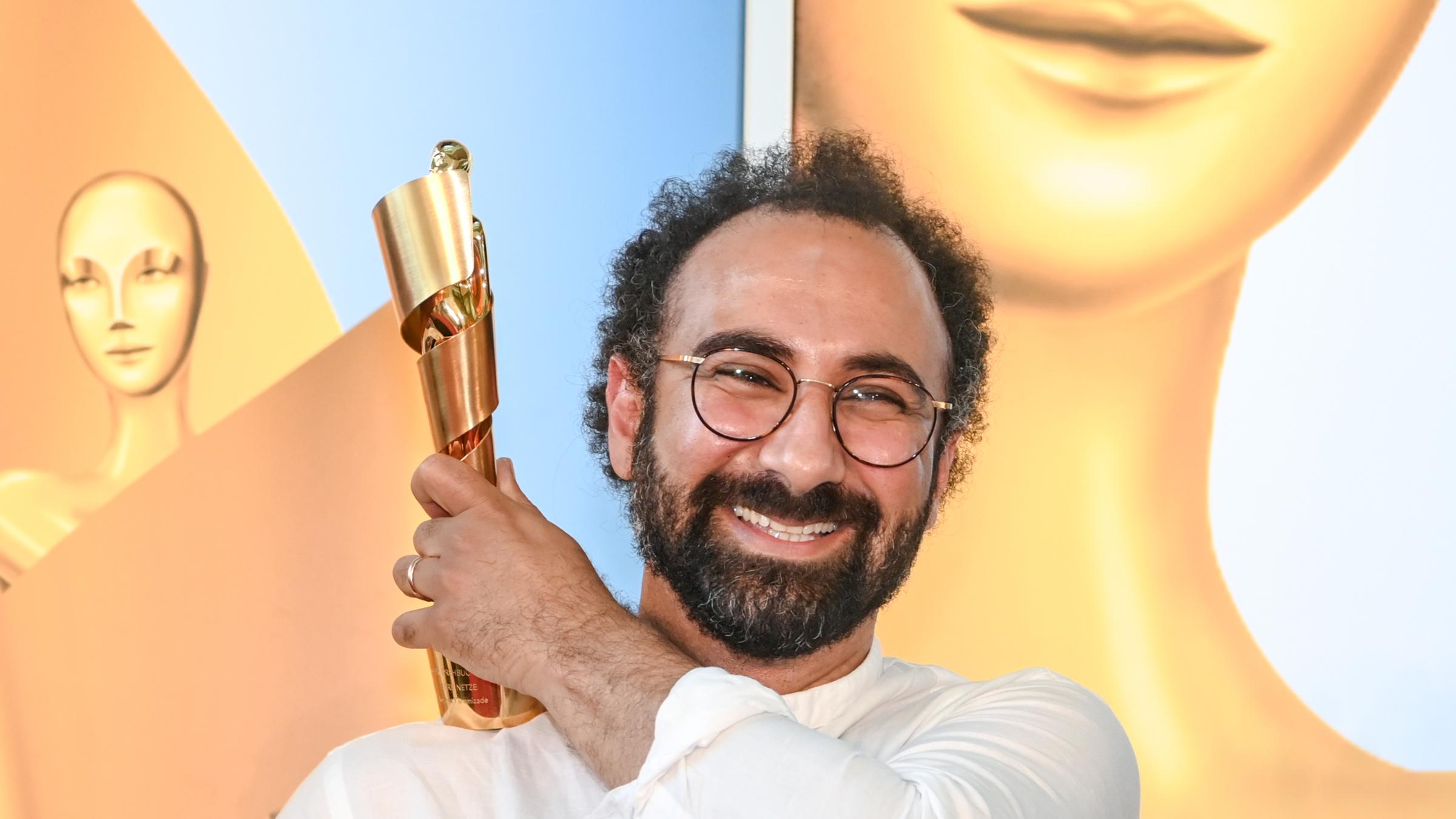 Der Autor Behrooz Karamizade(l.) präsentiert die Goldene Lola 2021 für sein Drehbuch "Leere Netze" mit der linken Hand auf seiner rechten Schulter und lacht in die Kamera.