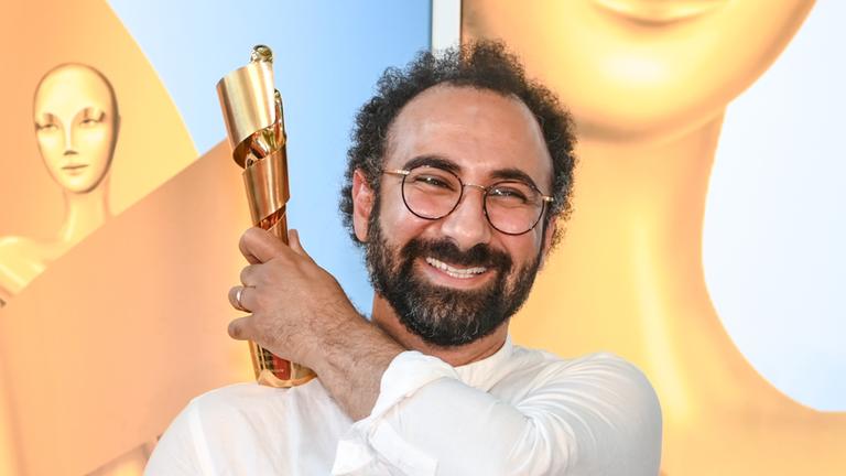 Der Autor Behrooz Karamizade(l.) präsentiert die Goldene Lola 2021 für sein Drehbuch "Leere Netze" mit der linken Hand auf seiner rechten Schulter und lacht in die Kamera.