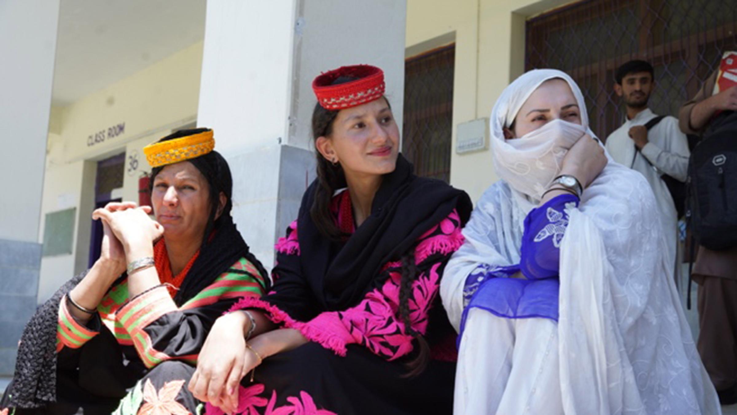 "Die Ungläubigen vom Hindukusch – Die bunte Kultur der Kalash" : Drei Frauen vom Volk der Kalasha sitzen vor einer Hauswand und blicken nach links. Sie tragen traditionelle Trachten.