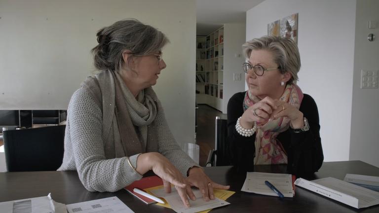 "Der Ast, auf dem ich sitze - Ein Steuerparadies in der Schweiz": Zwei Frauen mittleren Alters, beide tragen Brille, im Gespräch über Formulare, die auf einem schwarzen Tisch vor ihnen liegen. Im Hintergrund ist ein Sideboard an einer weißen, bilderlosen Wand und rechts ein langer Flur mit Bücherregalen zusehen.