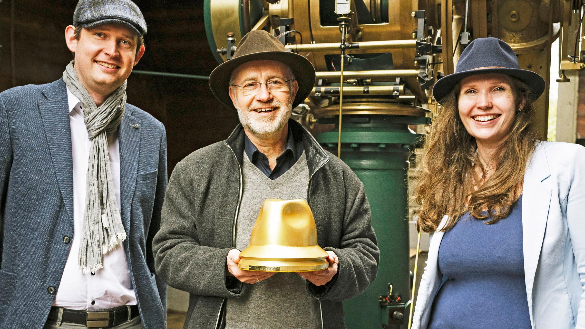 Harald Lesch mit Hut bei der Verleihung des Preises "Hutträger des Jahres 2021". Er hält die Trophäe in der Hand und  lächelt in die Kamera. Rechts neben ihm steht ein Vertreter, links eine Vertreterin des GDH e.V.; beide ebenfalls mit Kopfbedeckungen. 