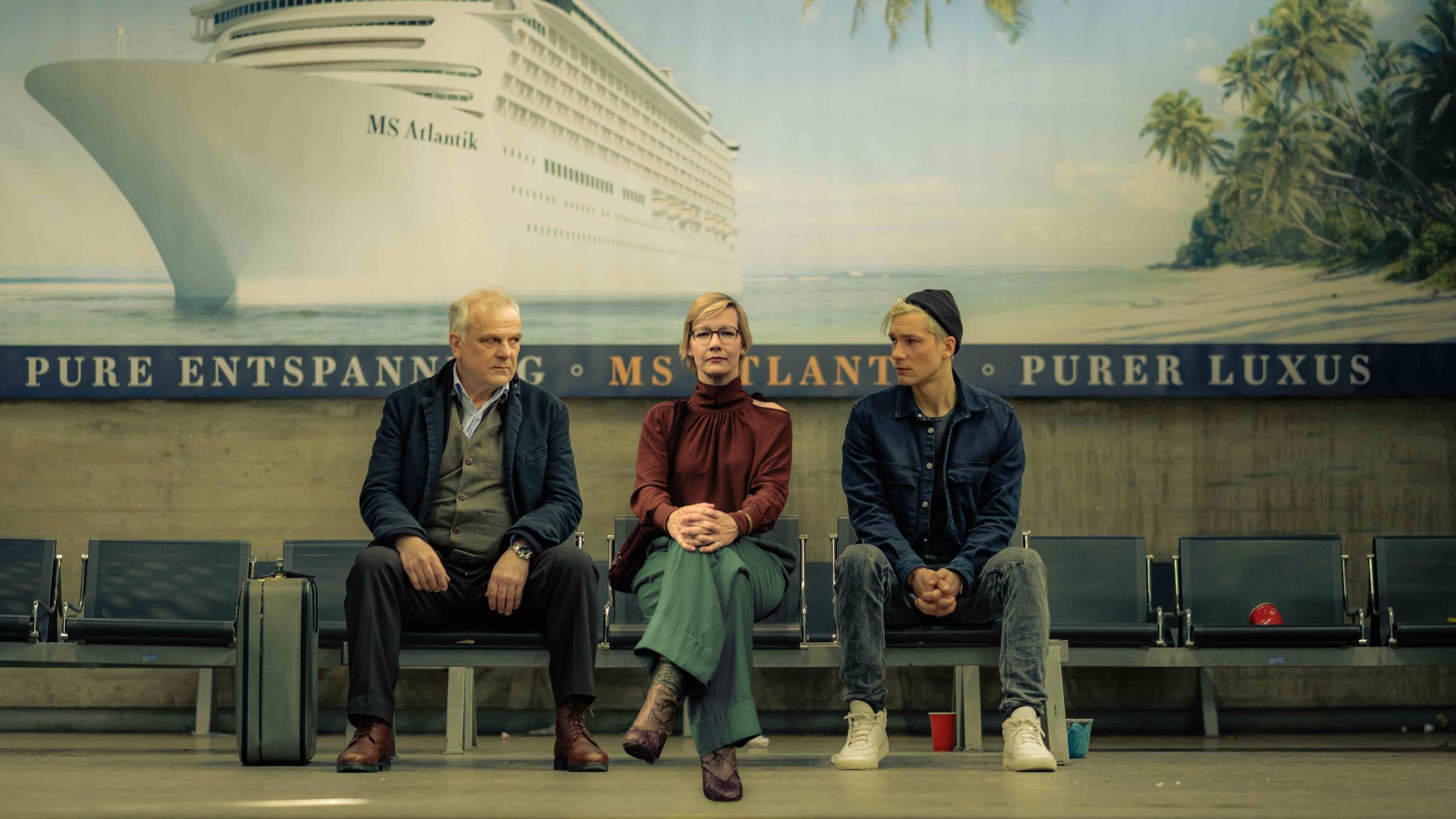 "Das schwarze Quadrat": Vincent Kowalski (Bernhard Schütz), Martha (Sandra Hüller) und Nils Forsberg (Jacob Matschenz) sitzen in einer Stuhlreihe nebeneinander. An der Wand hinter ihnen hängt ein großes Poster, das ein Kreuzfahrtschiff neben einem tropischen Sandstrand zeigt.