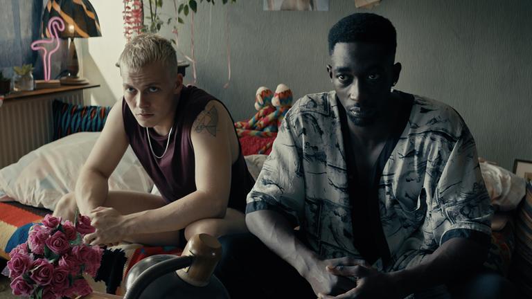 "TOUBAB": Dennis (Julius Nitschkoff)  und Babtou (Farba Dieng) sitzen nebeneinander auf einem Bett und blicken ernst in die Kamera.
