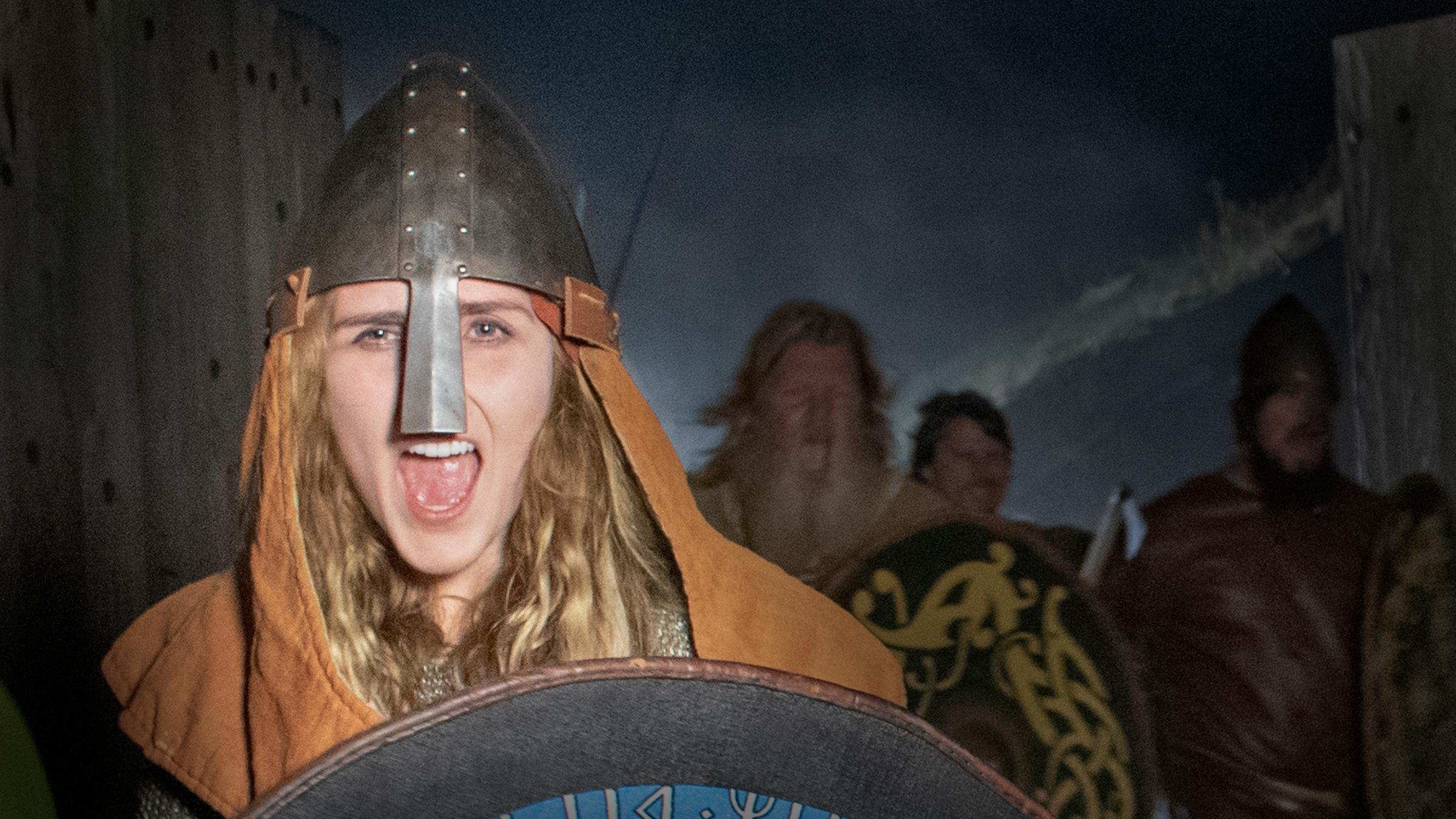 "Die Kriegerinnen der Wikinger": Die Kriegerin Signe (Moa Enqvist Stefansdotter) führt eine Wikingerhorde in den Kampf. Sie trägt einen Helm und ein Schild. Hinter ihr im Halbdunkel drei männliche Krieger,