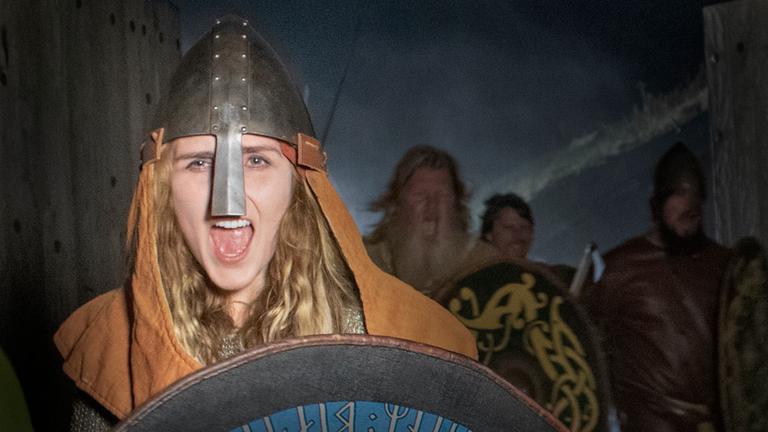 "Die Kriegerinnen der Wikinger": Die Kriegerin Signe (Moa Enqvist Stefansdotter) führt eine Wikingerhorde in den Kampf. Sie trägt einen Helm und ein Schild. Hinter ihr im Halbdunkel drei männliche Krieger,