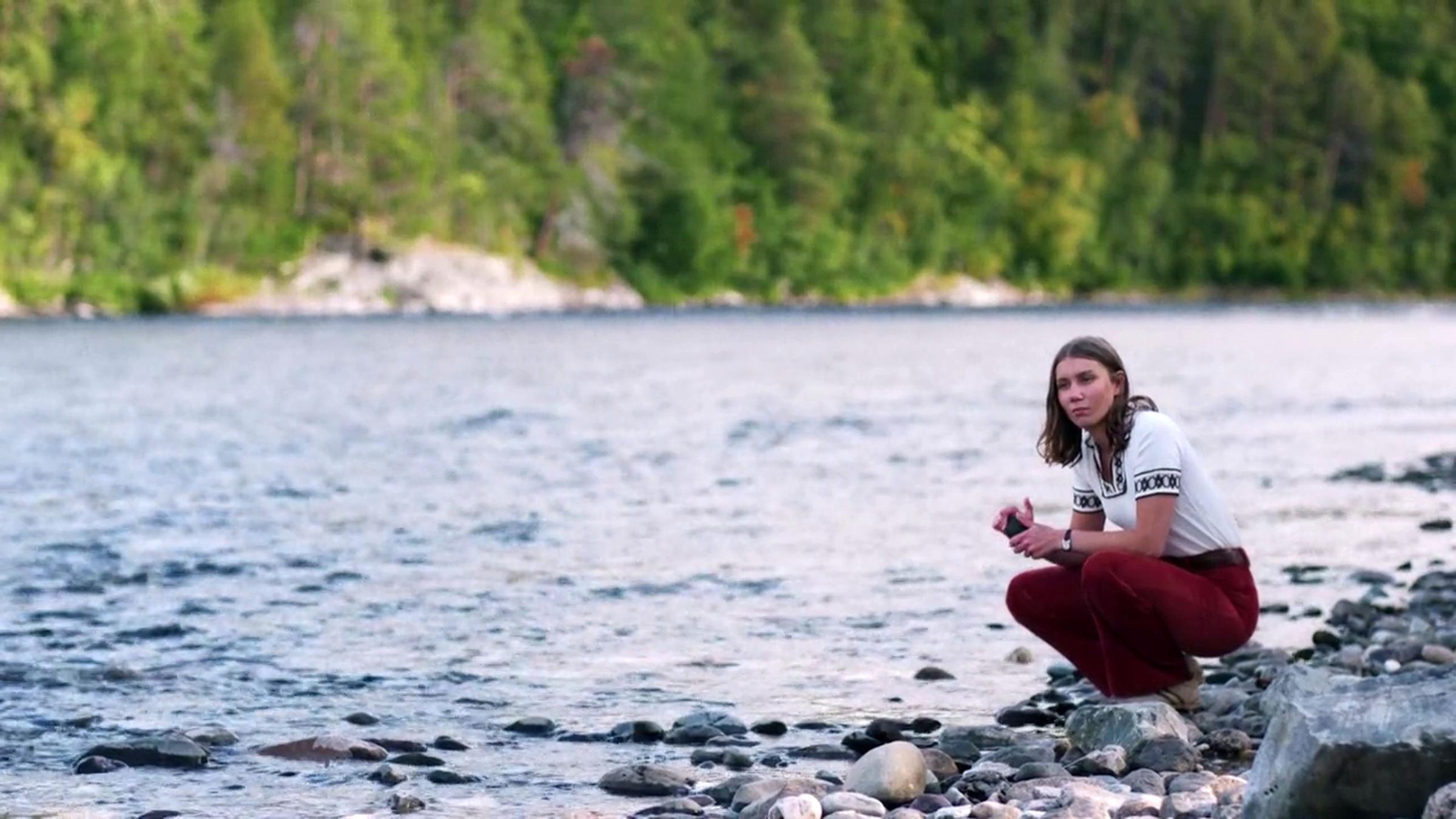 "Ellos eatnu – Let the River Flow": Ester (Ella Marie Hætta Isaksen) hockt am steinigen Ufer eines kleinen Flusses. Im Hintergrund ist das gegenüber liegende, bewaldete Flußufer zu sehen.