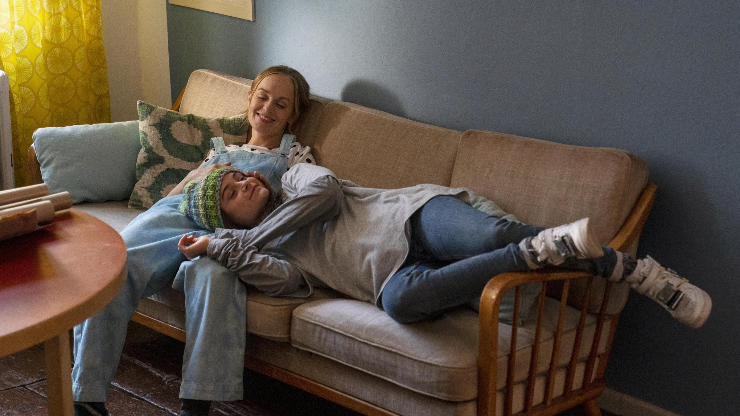 "Sweet Disaster": Frida (Friedrike Kempter) sitzt lächelnd auf einem Sofa. Neben ihr liegt Yolanda (Lena Urzendowsky) mit ihrem Kopf in Lenas Schoß. Sie hat die Augen geschlossen und eine Mütze auf em Kopf