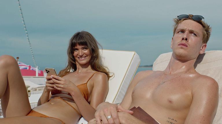 "Triangle of Sadness": Carl (Harris Dickinson) und Yaya ( Charlbi Dean Kriek) sitzen nebeneinander auf Matratzen am Strand zund schauen in die Kamera.