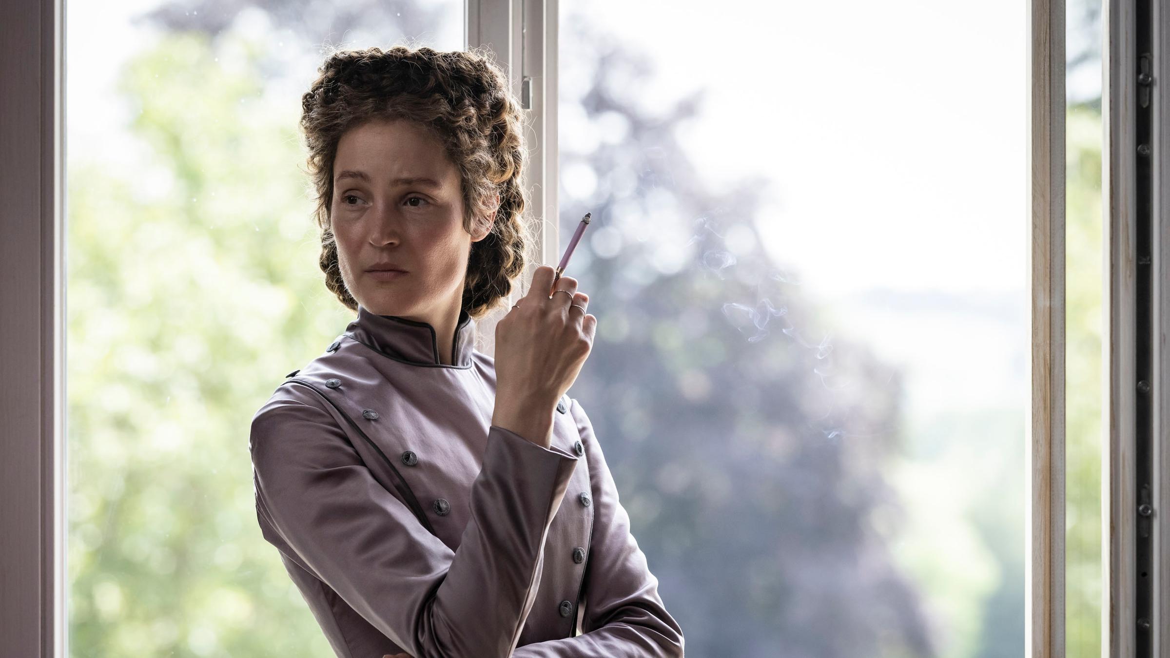 "Corsage": Kaiserin Elisabeth (Vicky Krieps) steht mit einer Zigarette in der rechten Hand am Fenster und blickt mit nachdenklichem Gesichtsausdruck übr ihre rechte Schulter.