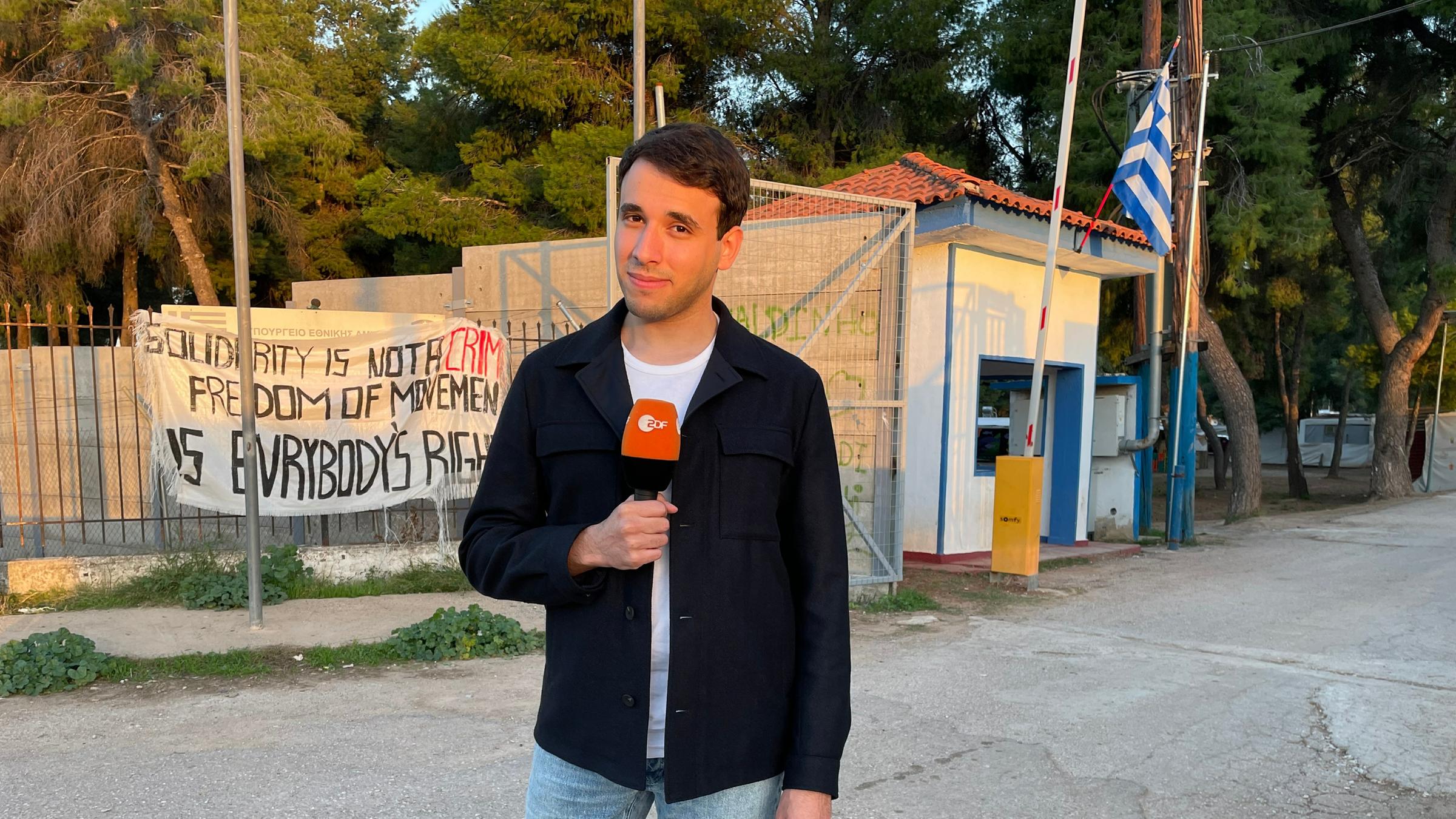"logo! extra: Kinder auf der Flucht – Wer kümmert sich um ihre Rechte?“: Sherif Rizkallah steht mit ein em Mikrofon in der Rechten Hand auf der Straße vor dem Eingang zu einem Flüchtlingslager auf der grichischen Insel Lesbos und blickt in die Kamera.