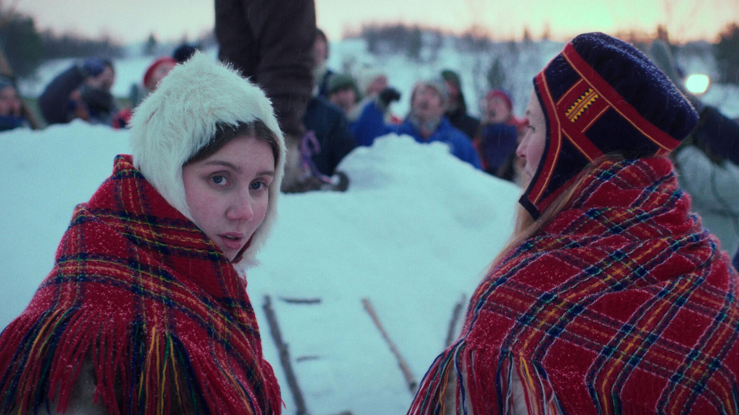 „Ellos eatnu - Let the River Flow“ („Ellos eatnu – La elva leve“) : Zwei samische Frauen stehen nebeneinander vor einem Schneehügel. Die linke blickt sich nach hinten um. Im Hintergrund ist unscharf eine Menschenansammlung zu erkennen.